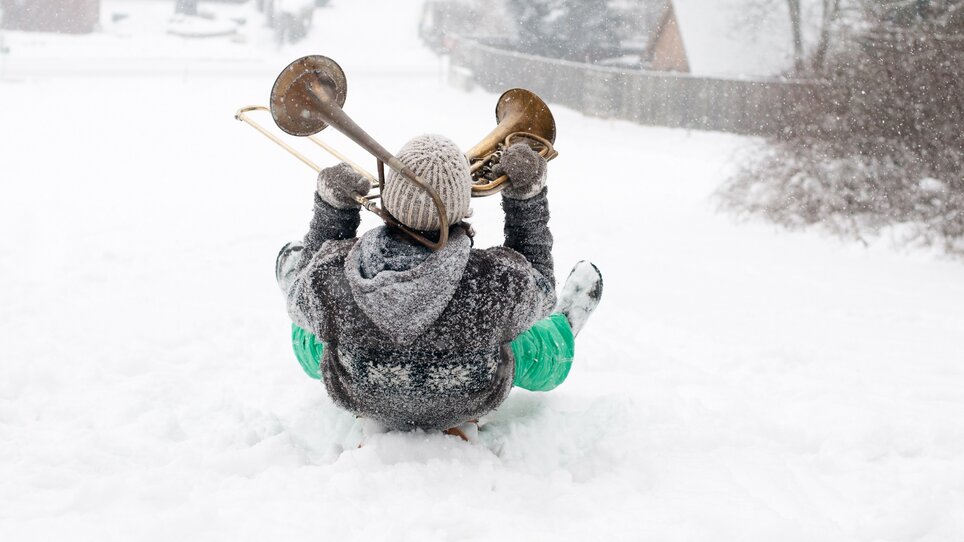 Eine Person sitzt auf einem Schlitten in verschneiter Landschaft und spielt Posaune. | © Steirisches Volksliedwerk - Unterrainer