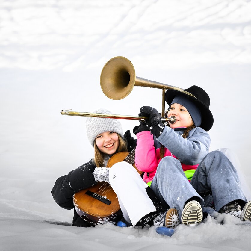 Zwei Kinder im Schnee, eines mit Trompete und das andere mit Gitarre, fröhlich auf einem Schlitten. | © Ulrike Rauch