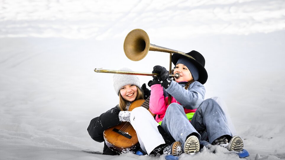 Zwei Kinder im Schnee, eines mit Trompete und das andere mit Gitarre, fröhlich auf einem Schlitten. | © Ulrike Rauch