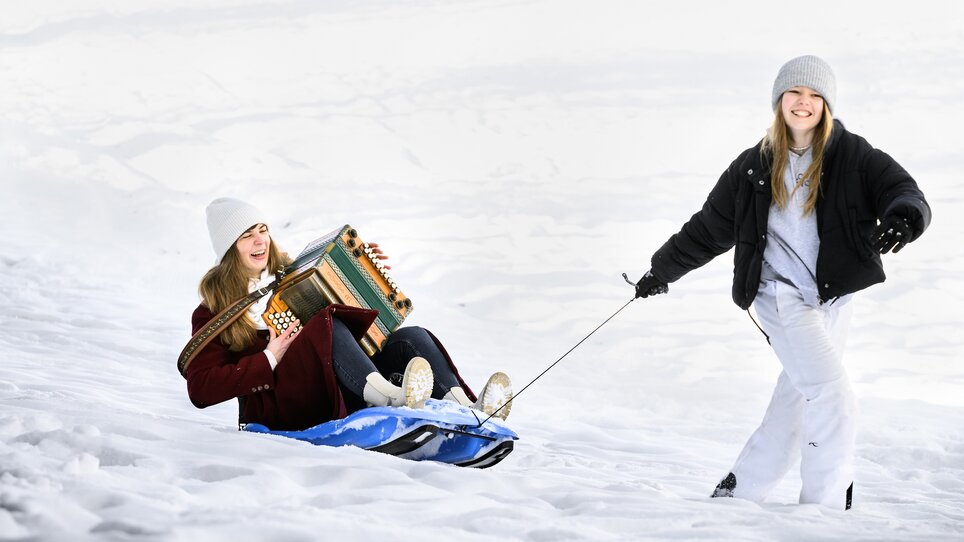 Zwei Mädchen im Schnee, eins spielt Melodika auf einem Schlitten, das andere zieht. | © Ulrike Rauch
