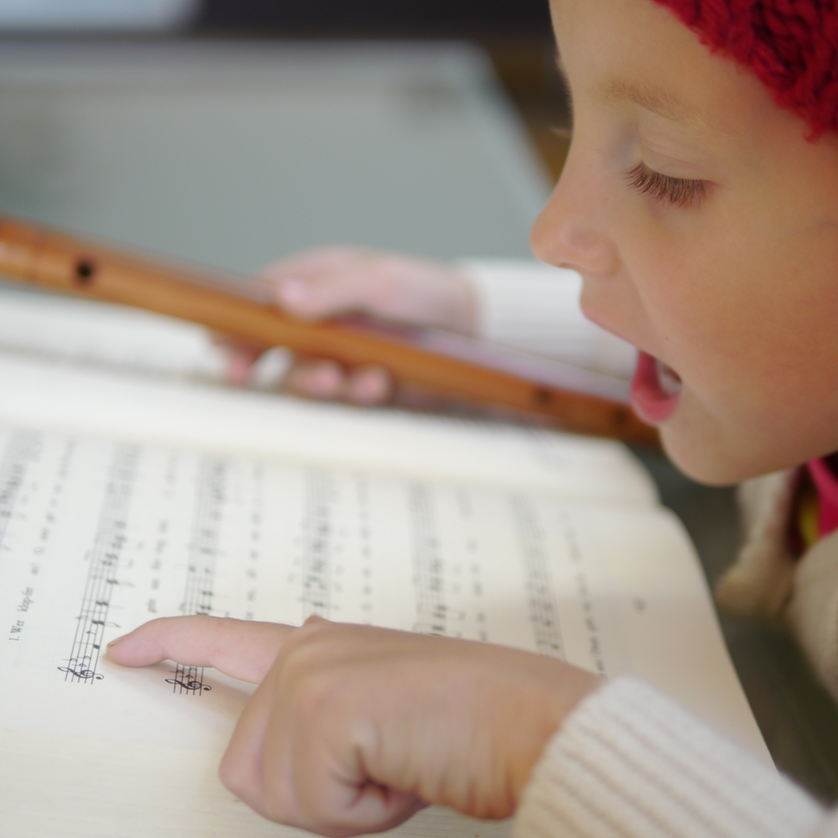 Ein Mädchen mit roter Mütze singt und spielt Flöte, während sie auf Notenblätter schaut. | © Steirisches Volksliedwerk