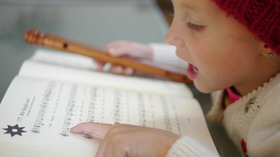 Ein Mädchen mit roter Mütze singt und spielt Flöte, während sie auf Notenblätter schaut. | © Steirisches Volksliedwerk