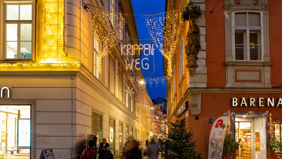 Beleuchtete Straßen mit 'Krippen Weg' Schild in Graz während der Abenddämmerung. | © Graz Tourismus-Harry Schiffer