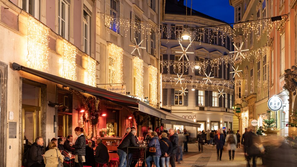 Belebte Stempfergasse in Graz, festlich mit Lichtern und Menschen, die Getränke genießen. | © Graz Tourismus-Harry Schiffer