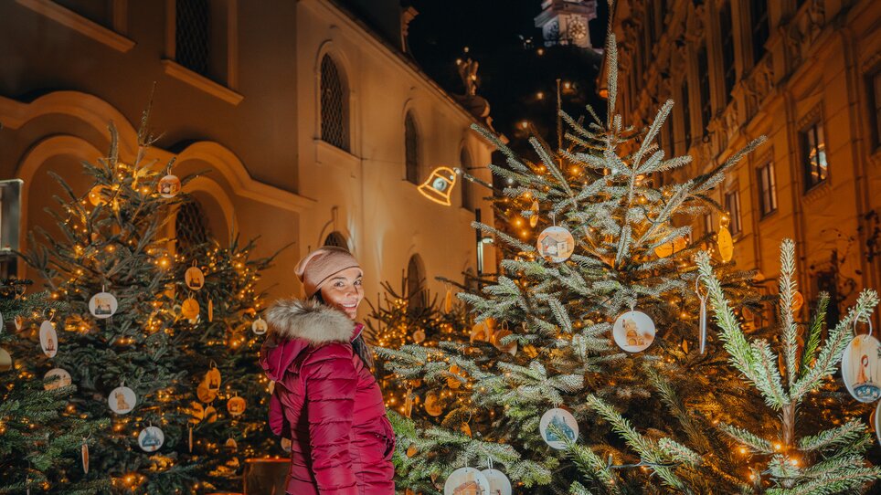 A woman in a red jacket stands among decorated Christmas trees in Graz, with the clock tower in the background. | © Mias Photoart 