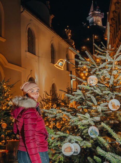 Eine Frau in einer roten Jacke steht zwischen geschmückten Weihnachtsbäumen in Graz, mit dem Uhrturm im Hintergrund. | © Mias Photoart 
