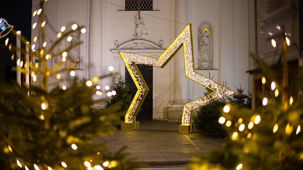 A large illuminated star stands in front of a church in Graz, surrounded by festive decorated fir trees. | © Graz Tourismus - Harry Schiffer