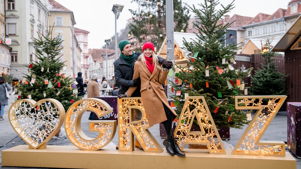 Paar posiert vor beleuchteter Graz-Dekoration im Weihnachtsmarkt. | © Graz Tourismus - Mias Photoart