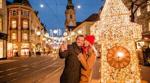Couple takes a selfie in front of the illuminated clock tower in Graz. | © Graz Tourismus - Mias Photoart