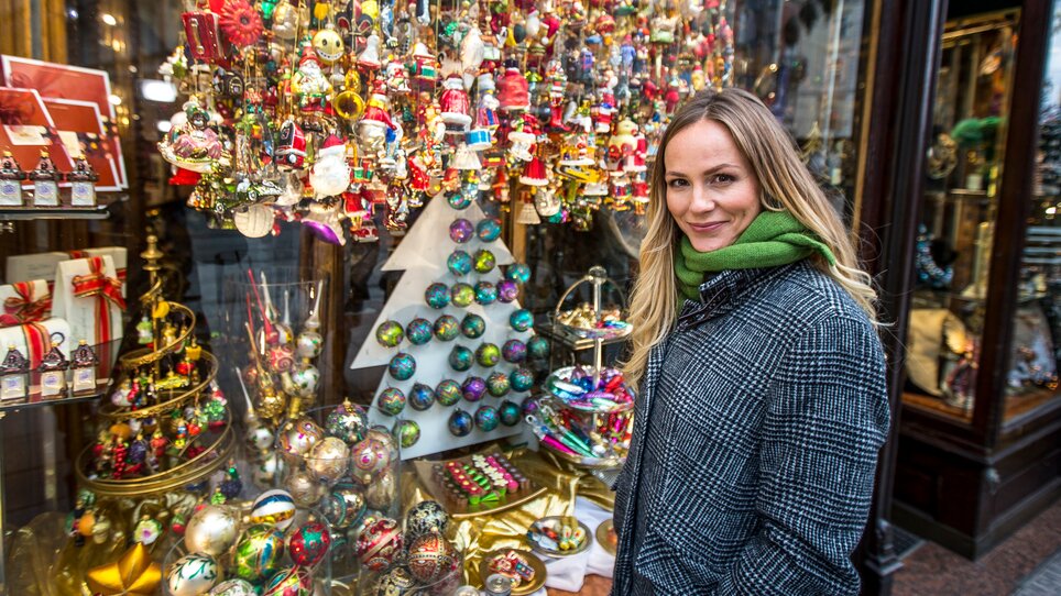 A woman stands in front of a shop window decorated for Christmas in Graz. The window displays colorful ornaments and Christmas figures. | © Graz Tourismus - Tom Lamm
