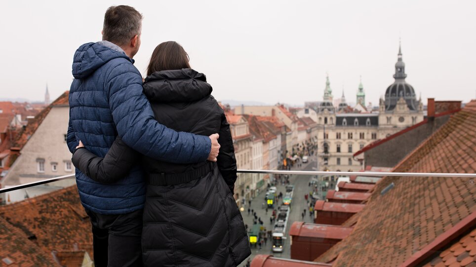 Coppia che guarda da una terrazza il panorama dei tetti di Graz e il municipio. | © Graz Tourismus - Katja Stadler