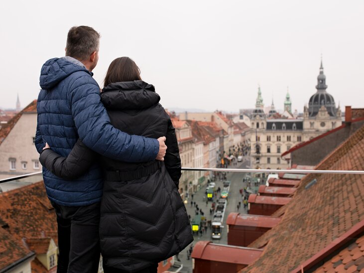 Paar schaut von einer Terrasse auf die Dachlandschaft von Graz und das Rathaus. | © Graz Tourismus - Katja Stadler