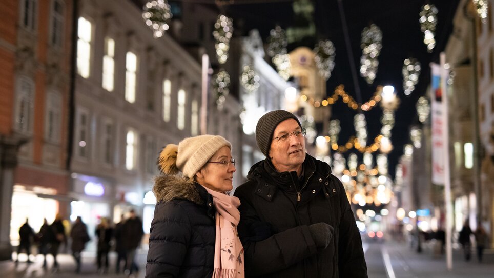 A couple walks under festive lighting in Graz, the parish church is visible. | © Graz Tourismus - Katja Stadler