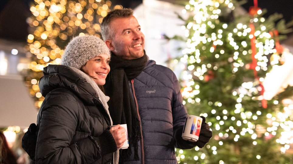 A couple in winter clothes with mugs in front of a festively lit Christmas tree. | © Graz Tourismus - Katja Stadler
