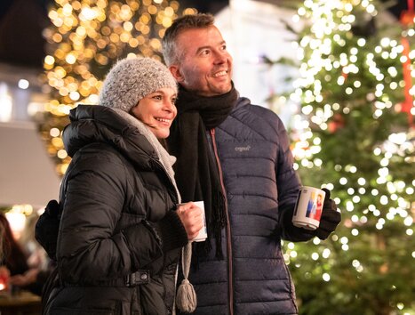 A couple in winter clothes with mugs in front of a festively lit Christmas tree. | © Graz Tourismus - Katja Stadler