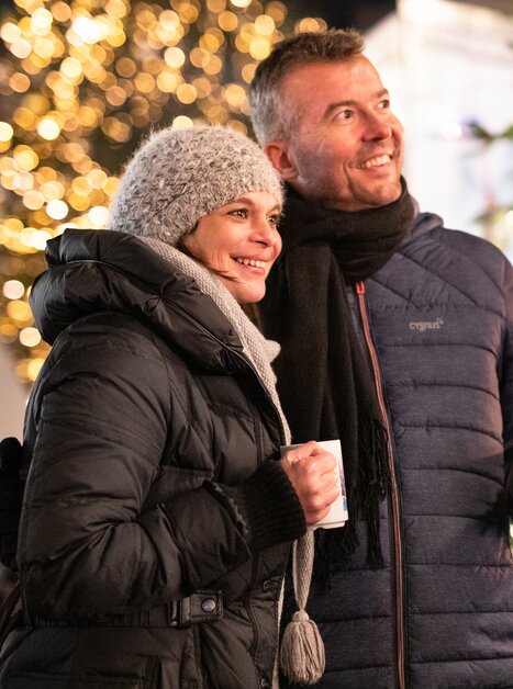 A couple in winter clothes with mugs in front of a festively lit Christmas tree. | © Graz Tourismus - Katja Stadler