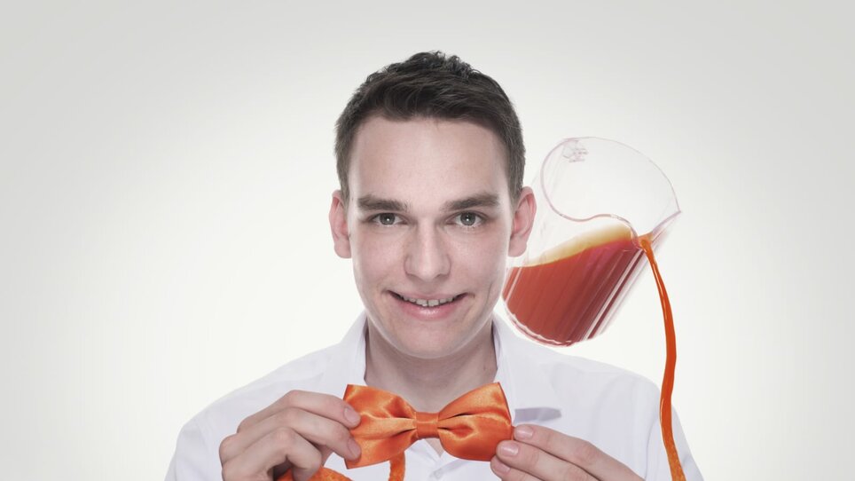 A man holds an orange bow tie and smiles as a jug of juice floats behind him. | © Zauberer Patrick