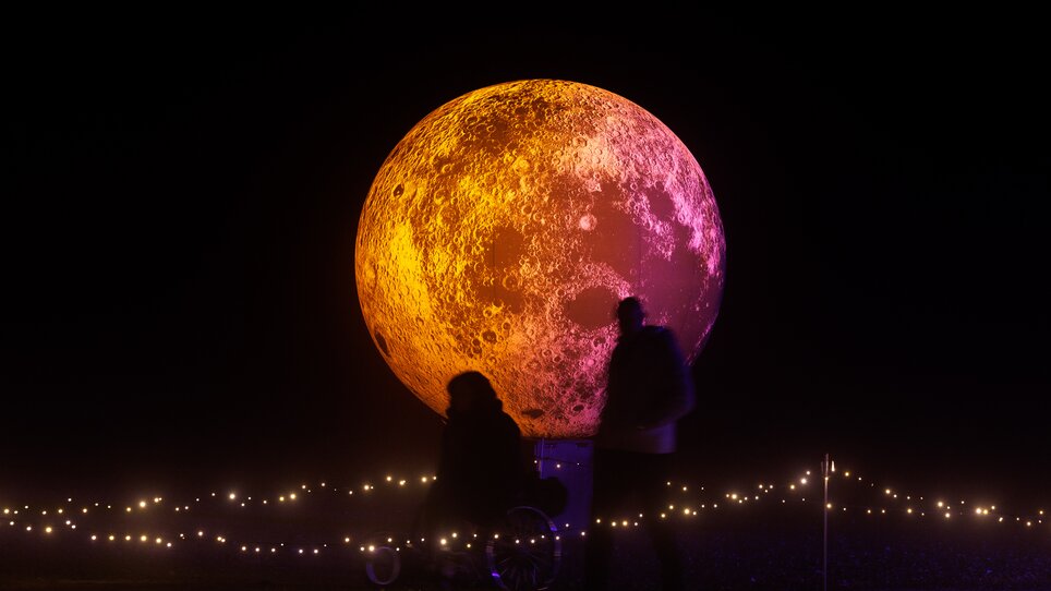 Una grande scultura di luna illuminata di notte con persone. | © Erwin Scheriau