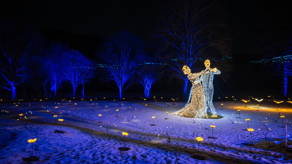 Bellissima installazione luminosa di una coppia che danza in un parco, circondata da alberi illuminati e un terreno innevato. | © Lumagica Frohnleiten