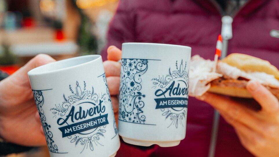 Zwei Tassen mit 'Advent am Eisernen Tor' und ein belegtes Brötchen in Händen. | © Mias Photoart