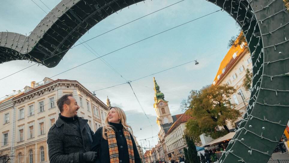 Zwei Personen im Winter auf dem Adventmarkt in Graz, mit der Stadtpfarrkirche im Hintergrund. | © Mias Photoart