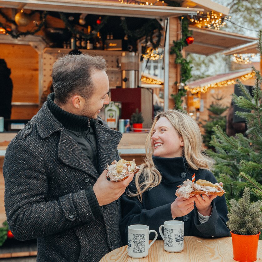 Advent in Graz I Adventmarkt am Eisernen Tor | © Mias Photoart Paar am Adventmarkt mit Snacks und Tassen in der Hand, umgeben von Lichtern und Weihnachtsbaum. | © Mias Photoart