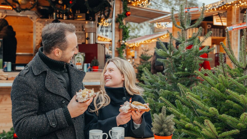 Paar am Adventmarkt mit Snacks und Tassen in der Hand, umgeben von Lichtern und Weihnachtsbaum. | © Mias Photoart