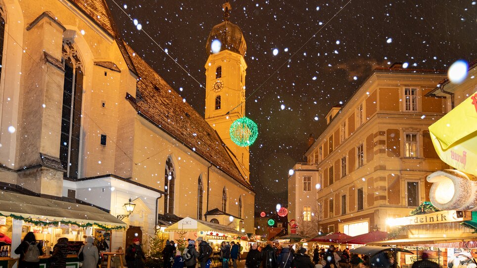 Schnee fällt über den Weihnachtsmarkt im Franziskanerviertel in Graz, mit beleuchteten Ständen und der beeindruckenden Architektur der Umgebung. | © Graz Tourismus-Harry Schiffer