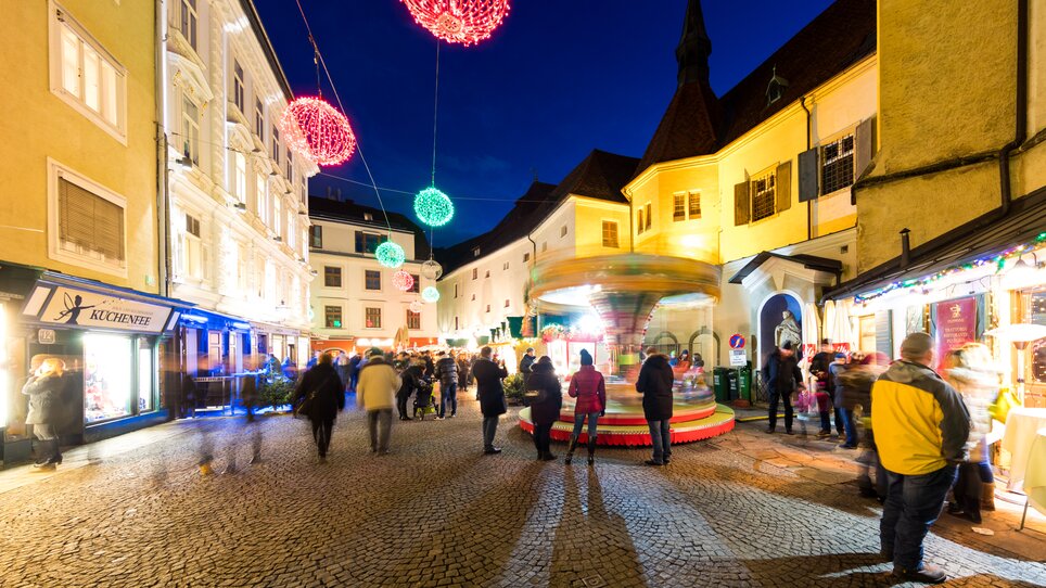 Belebte Straßen mit Lichtern, einem Karussell und Ständen beim Christkindlmarkt in Graz. | © Graz Tourismus - Harry Schiffer