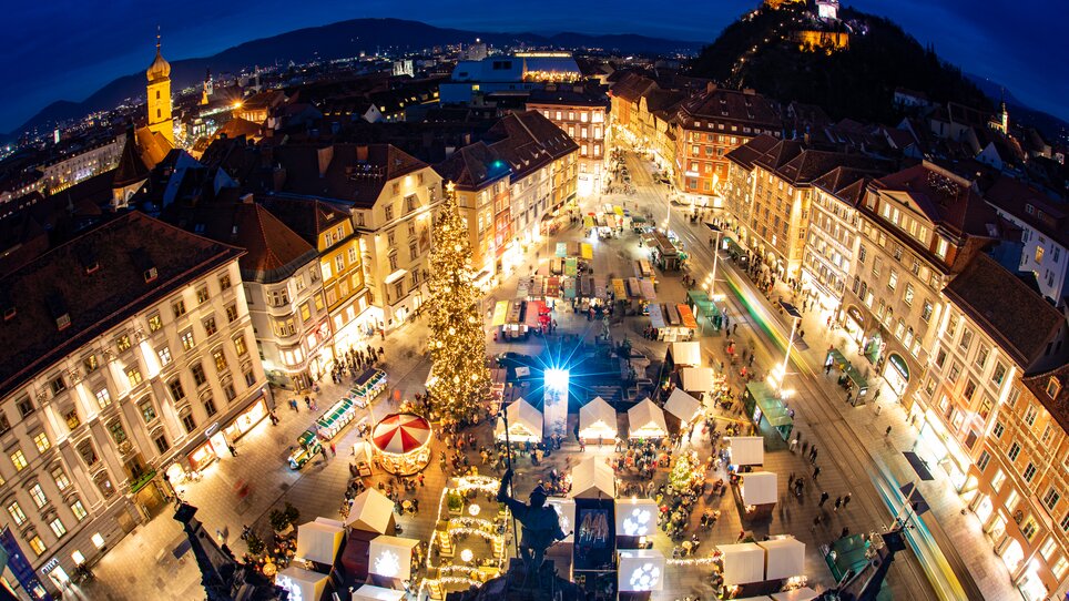 Illuminated Christmas market with stalls at the Hauptplatz of Graz, featuring the Graz Clock Tower in the background. | © Graz Tourismus - Harry Schiffer