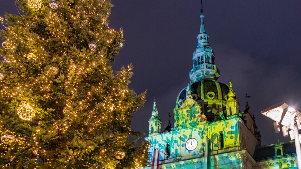 Illuminated Christmas tree and cheerful people in front of the town hall in Graz, with festive projection on the facade. | © achtzigzehn - Hinterleitner