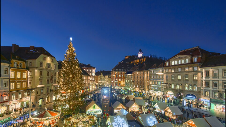 Blick auf den Weihnachtsmarkt in Graz mit festlich geschmückten Ständen, einem großen Weihnachtsbaum und dem Grazer Uhrturm im Hintergrund. | © Region Graz - Robert Maybach (RBMH) 