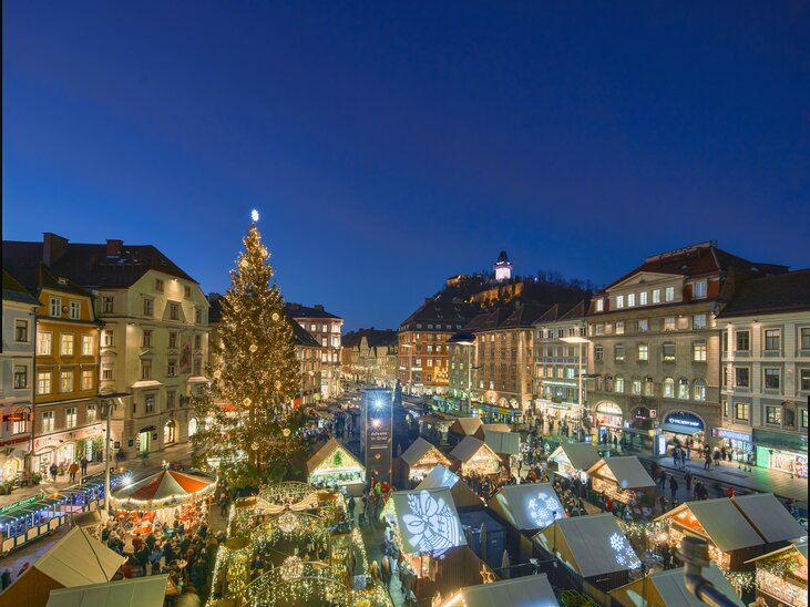 View of the Christmas market in Graz with festively decorated stalls, a large Christmas tree, and the Graz Clock Tower in the background. | © Region Graz - Robert Maybach (RBMH) 