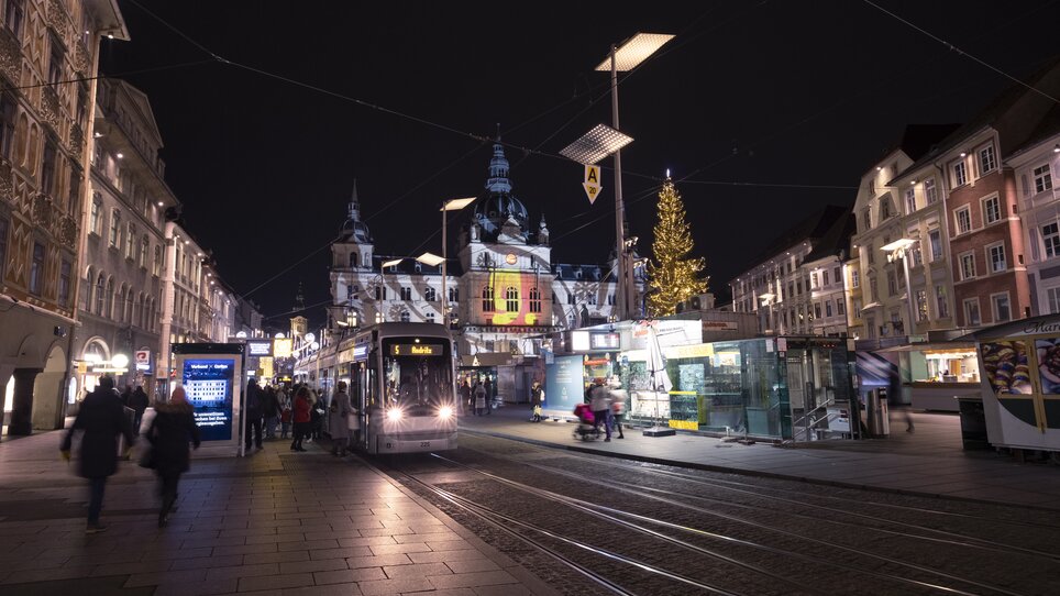 Nachtaufnahme eines Weihnachtsmarkts in Graz mit Straßenbahn und festlich beleuchtetem Rathaus. | © Vincent Croce
