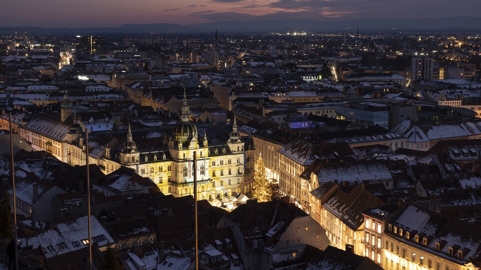 Blick auf Graz bei Nacht mit dem beleuchteten Rathaus und den Dächern der Stadt, umgeben von schneebedeckten Häusern. | © Vincent Croce