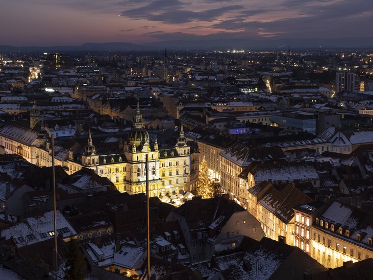 Blick auf Graz bei Nacht mit dem beleuchteten Rathaus und den Dächern der Stadt, umgeben von schneebedeckten Häusern. | © Vincent Croce