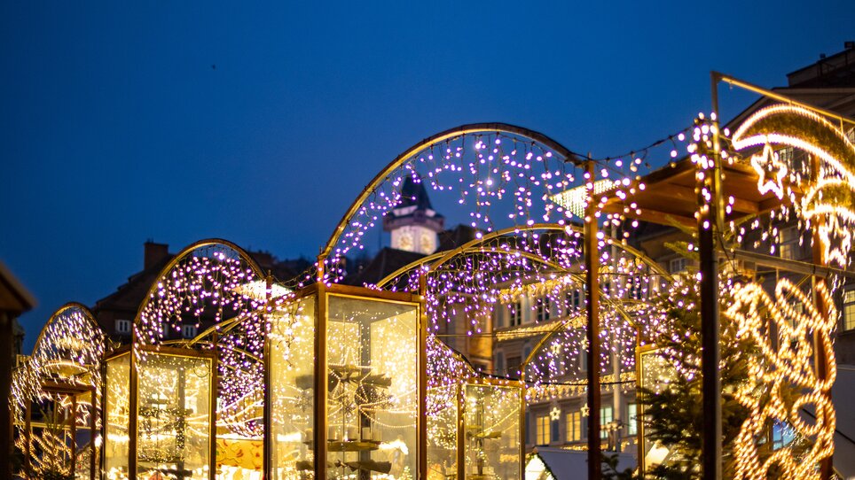 Beleuchteter Christkindlmarkt in Graz bei Nacht, mit Uhrturm im Hintergrund. | © Graz Tourismus - Harry Schiffer