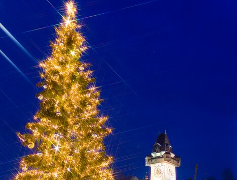 Ein festlich beleuchteter Weihnachtsbaum mit dem Grazer Uhrturm im Hintergrund, hell erleuchtet gegen den Abendhimmel. | © Graz Tourismus - Harry Schiffer