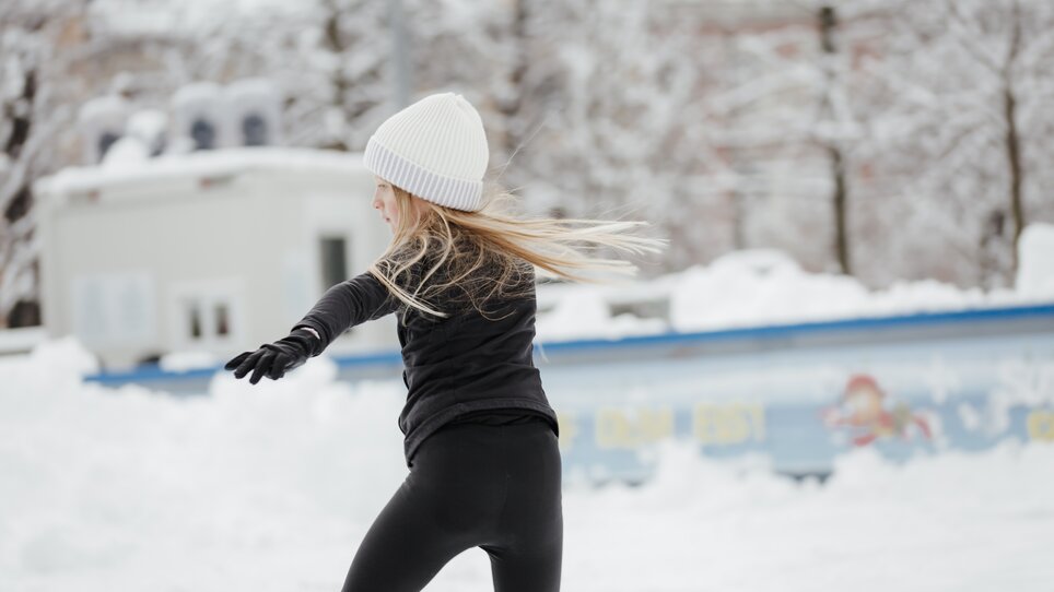 Ein Mädchen mit langen blonden Haaren und einer weißen Mütze, das auf einem Eislaufplatz im Winter Schlittschuh läuft. | © Daniel Kindler