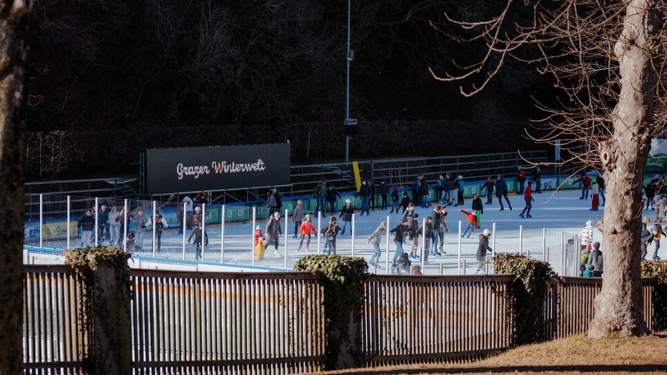 Menschen beim Eislaufen auf dem Eislaufplatz Grazer Winterwelt, umgeben von winterlicher Landschaft. | © Daniel Kindler
