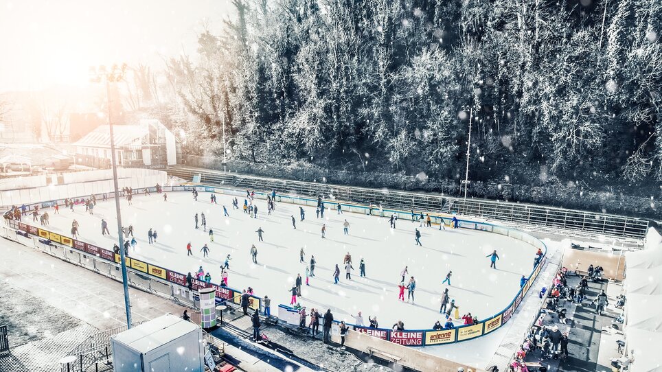 Menschen beim Eislaufen auf einem beleuchteten Eislaufplatz in Graz, umgeben von winterlicher Landschaft. | © Simon Möstl