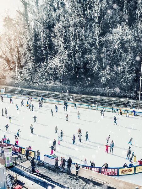Menschen beim Eislaufen auf einem beleuchteten Eislaufplatz in Graz, umgeben von winterlicher Landschaft. | © Simon Möstl
