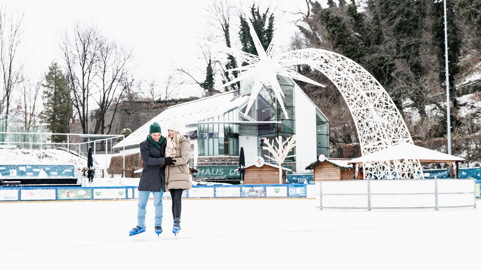 Ein Paar beim Eislaufen in der Winterwelt in Graz, mit Weihnachtsdekoration im Hintergrund. | © Graz Tourismus - Mias Photoart