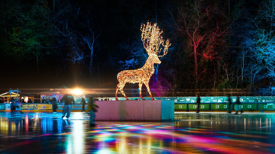 Beleuchtetes Rentier beim Eislaufplatz der Grazer Winterwelt bei Nacht, umgeben von bunten Lichtern und Besuchern. | © Leon Moik