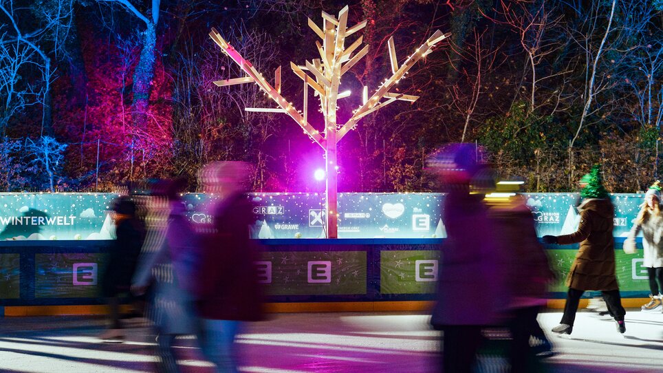Eisläufer auf einem beleuchteten Platz mit einem dekorierten Baum. | © Leon Moik