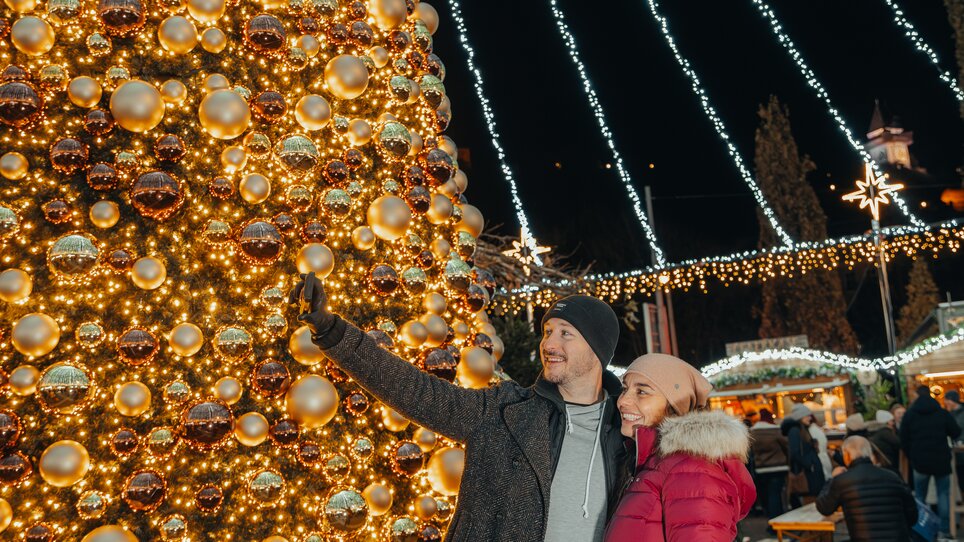 A couple poses in front of a beautifully decorated tree with lights and ornaments. | © Mias Photoart