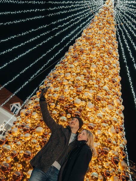 Ein Paar posiert vor einem großen, beleuchteten Weihnachtsbaum. | © Mias Photoart