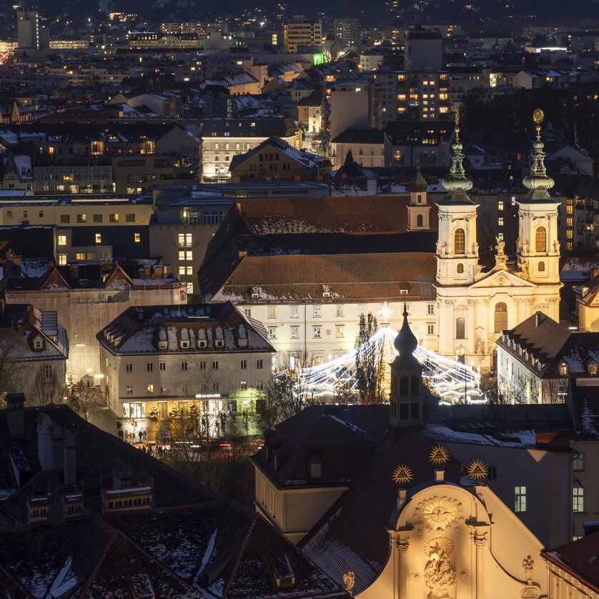 Beleuchtete Stadt Graz bei Nacht, mit Schnee auf den Dächern und dem Mariahilferplatz im Vordergrund. | © Vincent Croce