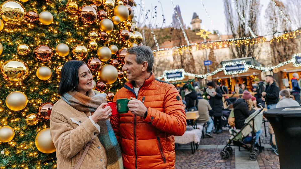 Couple smiling with mugs in front of Christmas tree and market in Graz. | © Graz Tourismus - Mias Photoart