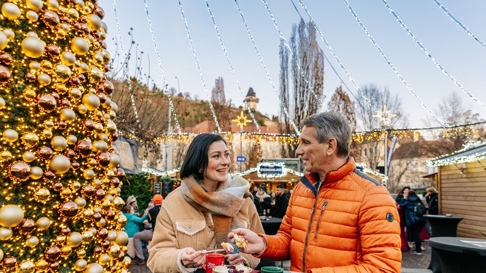 A couple enjoys snacks and drinks in front of a large Christmas tree at Mariahilferplatz in Graz. | © Graz Tourismus - Mias Photoart